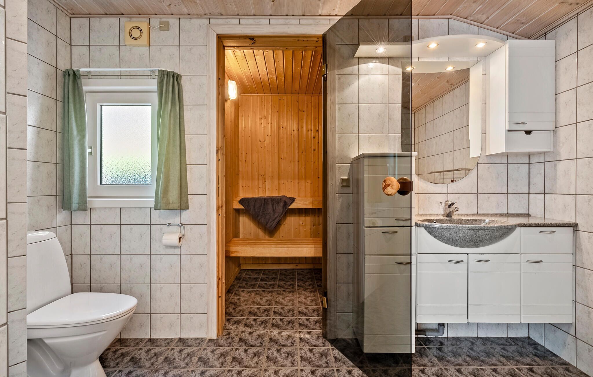 Et badeværelse med toilet, vask med kumme og en dør til en træsauna.