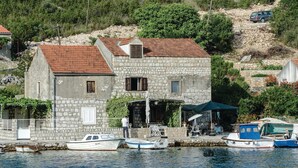 A stone building with a red-tiled roof, a small dock with boats, and a hillside in the background.