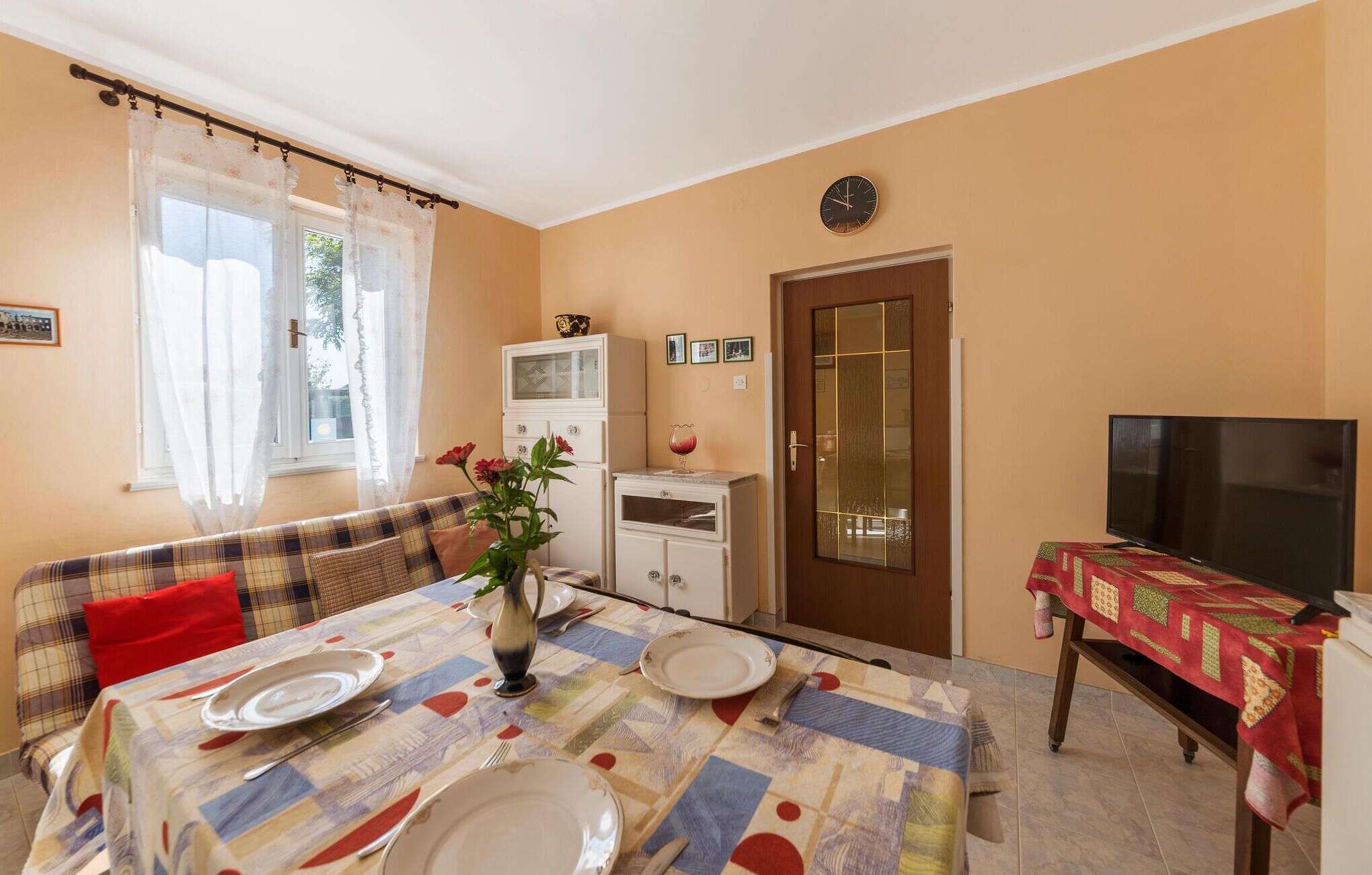 A dining area with a table set for two, a sofa, a television, and a cabinet.