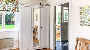 Una habitación con un armario blanco, una silla de madera, un ventanal con vistas a la vegetación y una puerta que da a otra habitación.