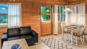 A cozy cabin interior with a wooden interior, a black leather sofa, a dining table with white chairs, and a window with a view of greenery. - 2 bedroom beach front home in Vallavik (Vallavik)