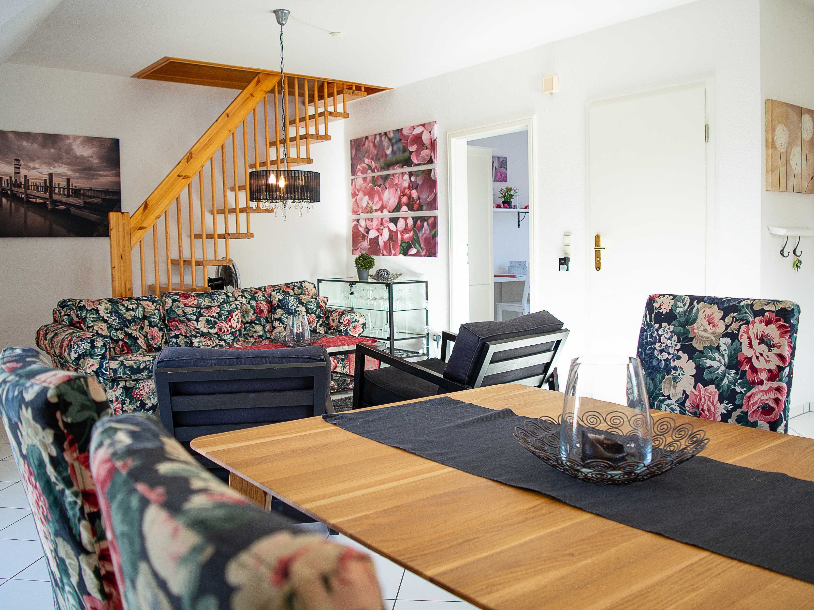 A modern living room with a wooden staircase, floral upholstery, a dining table with a glass centerpiece, and a view of the city through a window.