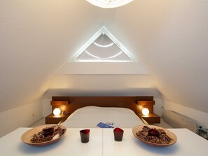 Ein Zimmer mit geneigter Decke, einem dreieckigen Oberlicht, einem Bett, Nachttischen mit Lampen und zwei Schalen auf einem Tisch.