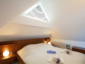 A bedroom with a skylight, a bedside table with a lamp, a clock, and a neatly made bed with towels and a brochure.