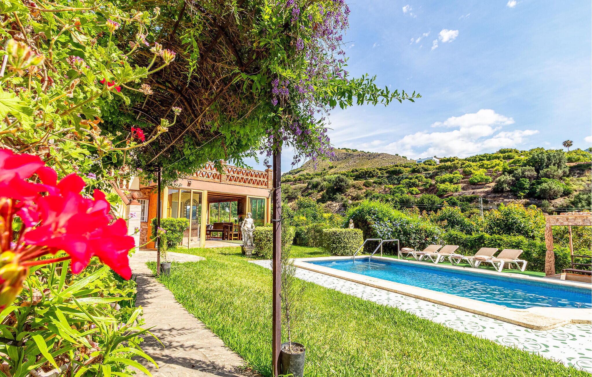 Un'area piscina con vista libera sulla vasca, un patio coperto e un giardino rigoglioso con diverse piante e fiori.