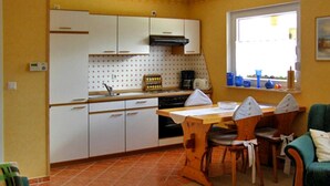A compact kitchen with white cabinets, a dining table, and chairs. A window provides natural light, and a green armchair is visible in the foreground. - Vacation cottage Ursula - Holiday cottage (Malchow)