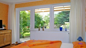 A bedroom with a large window, a wooden dresser, a bed with orange floral bedding, and a blue lamp. - Vacation cottage Ursula - Holiday cottage (Malchow)