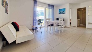 A modern living room with a white sofa, a dining table with chairs, and a view of the outdoors through a glass door.