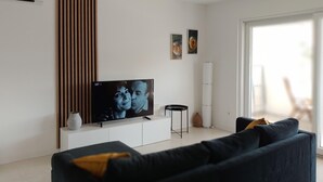 Una sala de estar moderna con un televisor de pantalla plana sobre un mueble blanco, un sofá oscuro con cojines amarillos y dos cuadros abstractos en la pared.