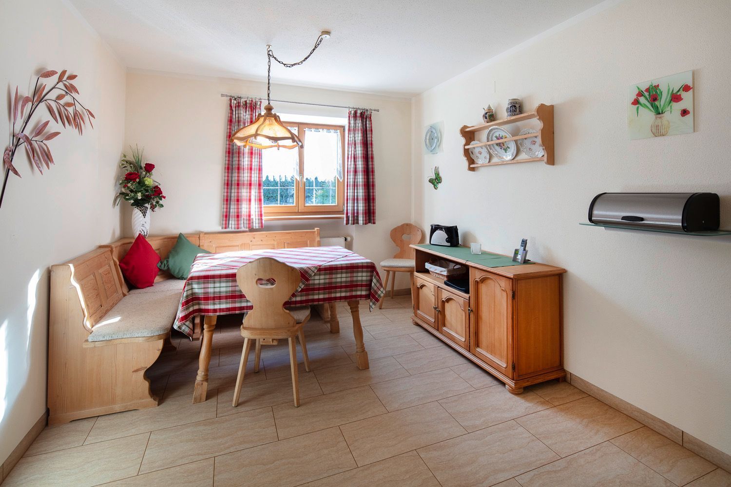 A cozy room with a bed, a wooden chair, a table, a desk, a shelf with dishes, a hanging light, and a window with curtains.