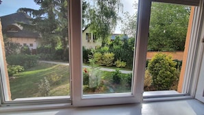 A window with a view of a well-maintained garden, a pathway, and a building with a sloped roof.