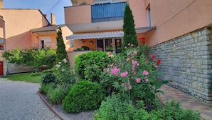 A residential building with a stone wall, a small garden with pink flowers, and a striped awning over the entrance.
