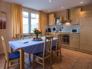 A dining area with a table set for four, a kitchen with basic appliances, and a window with curtains. - Three-room apartment type S6 with sea view - Strandstraße 24 (Karlshagen)
