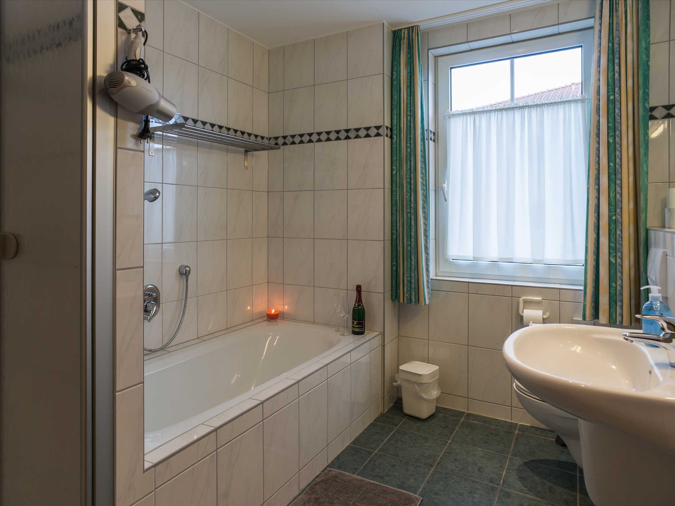 A bathroom with a bathtub, a window with curtains, a sink, and a trash bin.