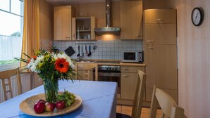 Een moderne keuken met houten kastjes, een magnetron, een oven en een spoelbak. Er staat een eettafel met een fruitschaal en een bloemenvaas.