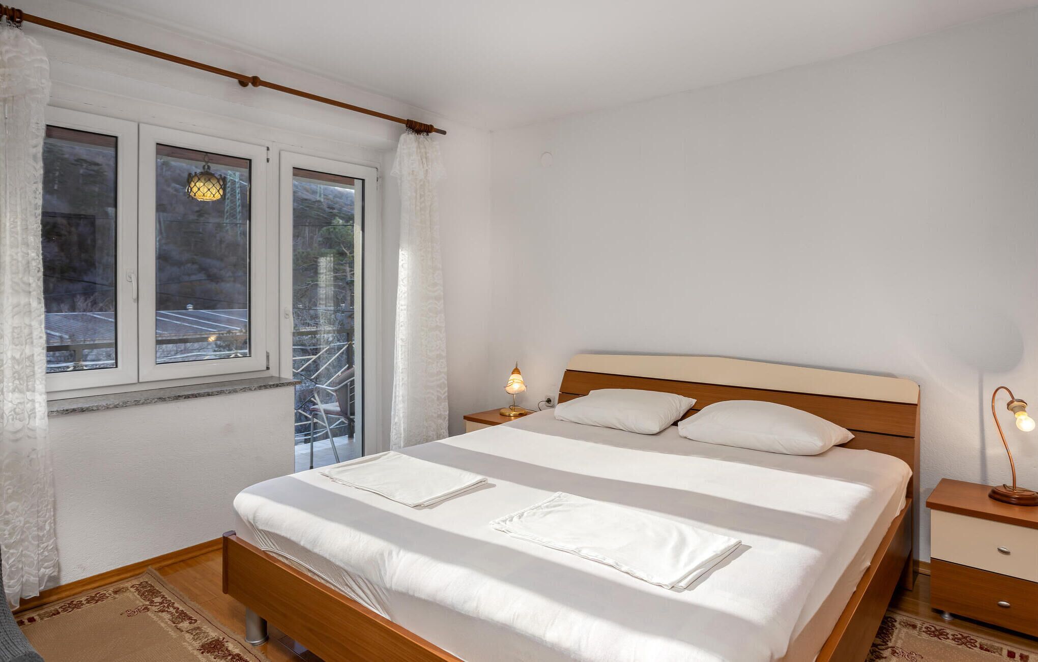 A bedroom with a wooden bed, white linens, a nightstand with a lamp, and a window with curtains.