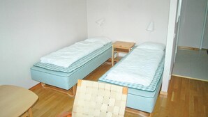Una habitación pequeña y sencilla con dos camas individuales, una mesita de noche de madera, una silla y una pequeña mesa.