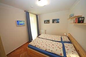 Une petite chambre avec deux lits simples, un téléviseur fixé au mur et une étagère remplie de livres.