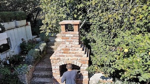Tiilistä rakennettu ulkouuni puutarhassa, ja sen edessä seisoo mies.