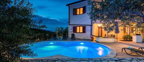 A two-story house with a swimming pool, stone wall, and outdoor furniture.