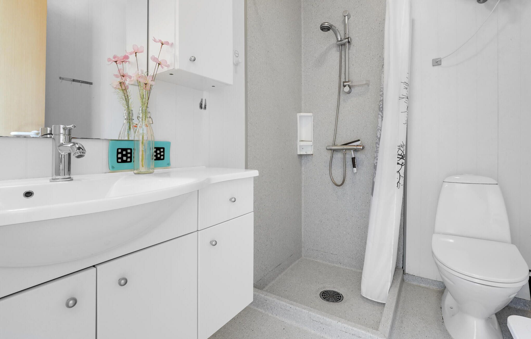 A modern bathroom with a white sink, a glass vase with pink flowers, a shower with a handheld showerhead, and a toilet.