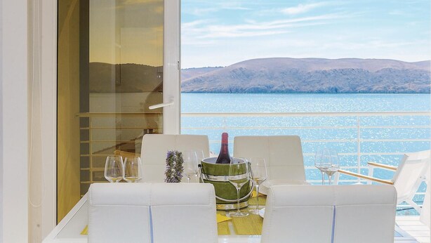 Une table à manger avec des chaises blanches, un seau vert et des verres à vin, donnant sur une étendue d’eau avec des montagnes en arrière-plan.