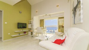 Un salon moderne avec un canapé blanc, un téléviseur à écran plat fixé au mur et un balcon offrant une vue sur la mer.