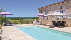 Zona de piscina con sillas de descanso y sombrillas, un edificio de piedra con contraventanas azules y vistas al campo.