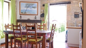Un comedor con una mesa dispuesta para cuatro, sillas de madera con cojines, un cuadro de un árbol junto al mar y una pared de piedra.