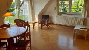 A spacious room with a dining table, chairs, a small table, and a window with a view of greenery. - Apartment 3 / o. r. - Ruegen-Fewo 47 (Putbus)