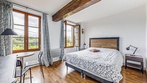 A bedroom with a large bed, a desk, and a chair. There are two windows with curtains, a radiator, and a wooden ceiling beam. - Appartement jolie vue rivière (Jaulgonne)