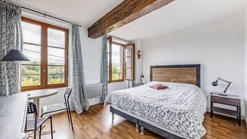 A bedroom with a large bed, a desk, and a chair. There are two windows with curtains, a radiator, and a wooden ceiling beam.