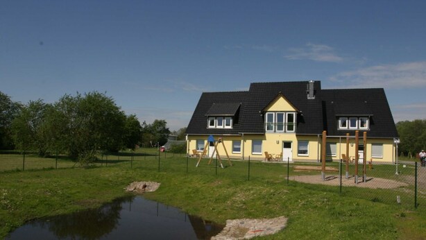 A yellow building with a black roof, surrounded by a fenced grassy area with a small pond.