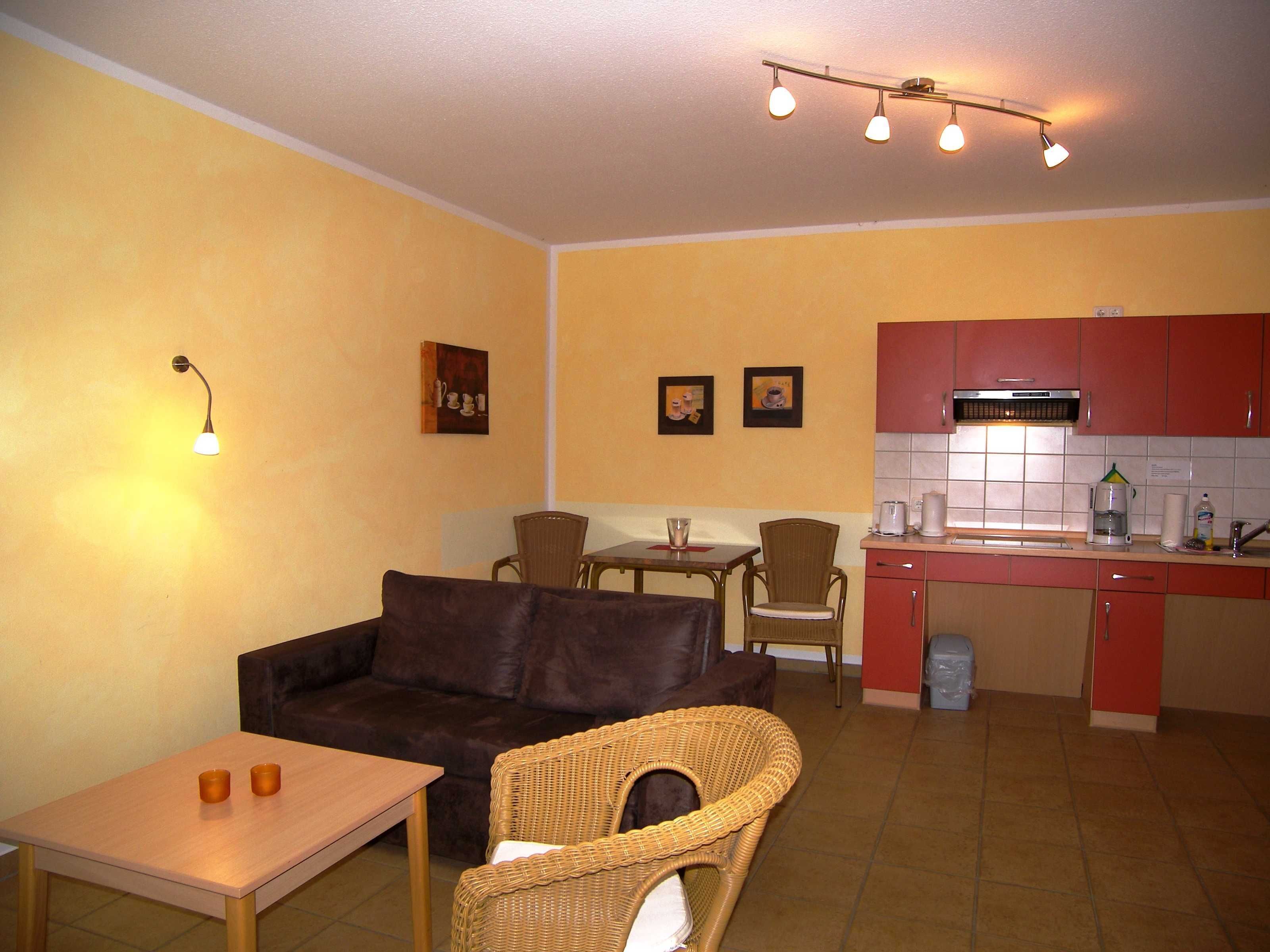 A compact living space with a sofa, dining table, chairs, a kitchenette, and a wall-mounted light fixture.