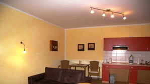 A compact living space with a sofa, dining table, chairs, a kitchenette, and a wall-mounted light fixture.