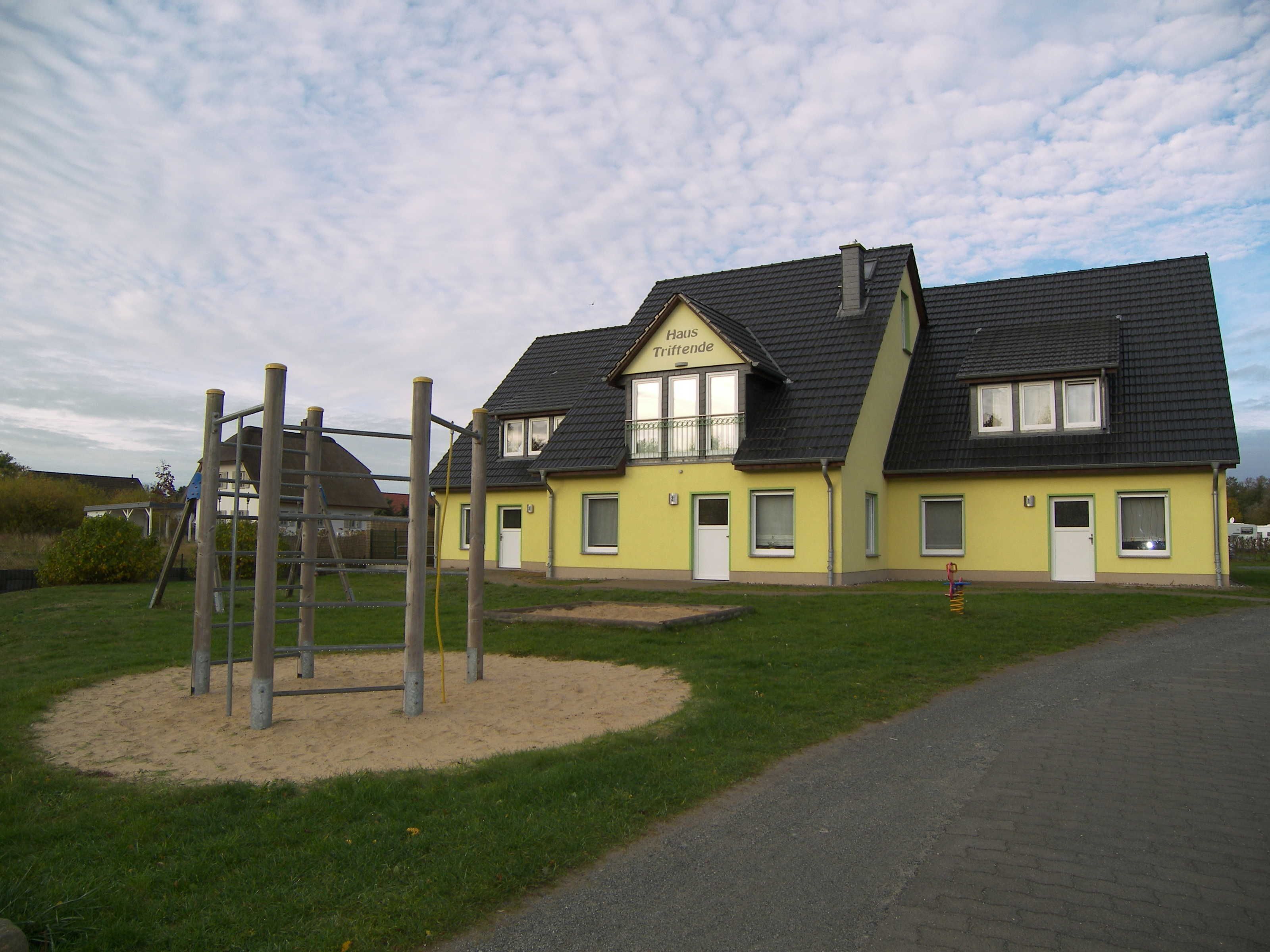 Ein zweistöckiges gelbes Gebäude mit dunklem Dach, ein Spielplatz mit einer hölzernen Kletterkonstruktion und einem gepflasterten Einfahrtsweg.