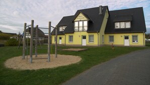 A two-story yellow building with a dark roof, a playground with a wooden climbing structure, and a paved driveway.