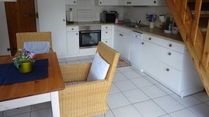 Una cocina con armarios blancos, una estufa y un lavabo. Se ve una silla de mimbre y una mesa con un florero.