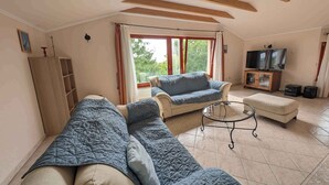 A spacious living room with a large sofa, a smaller armchair, a glass coffee table, and a television on a wooden cabinet. - Holiday home with pool and view to Lake Balaton for families (Balatonalmádi)