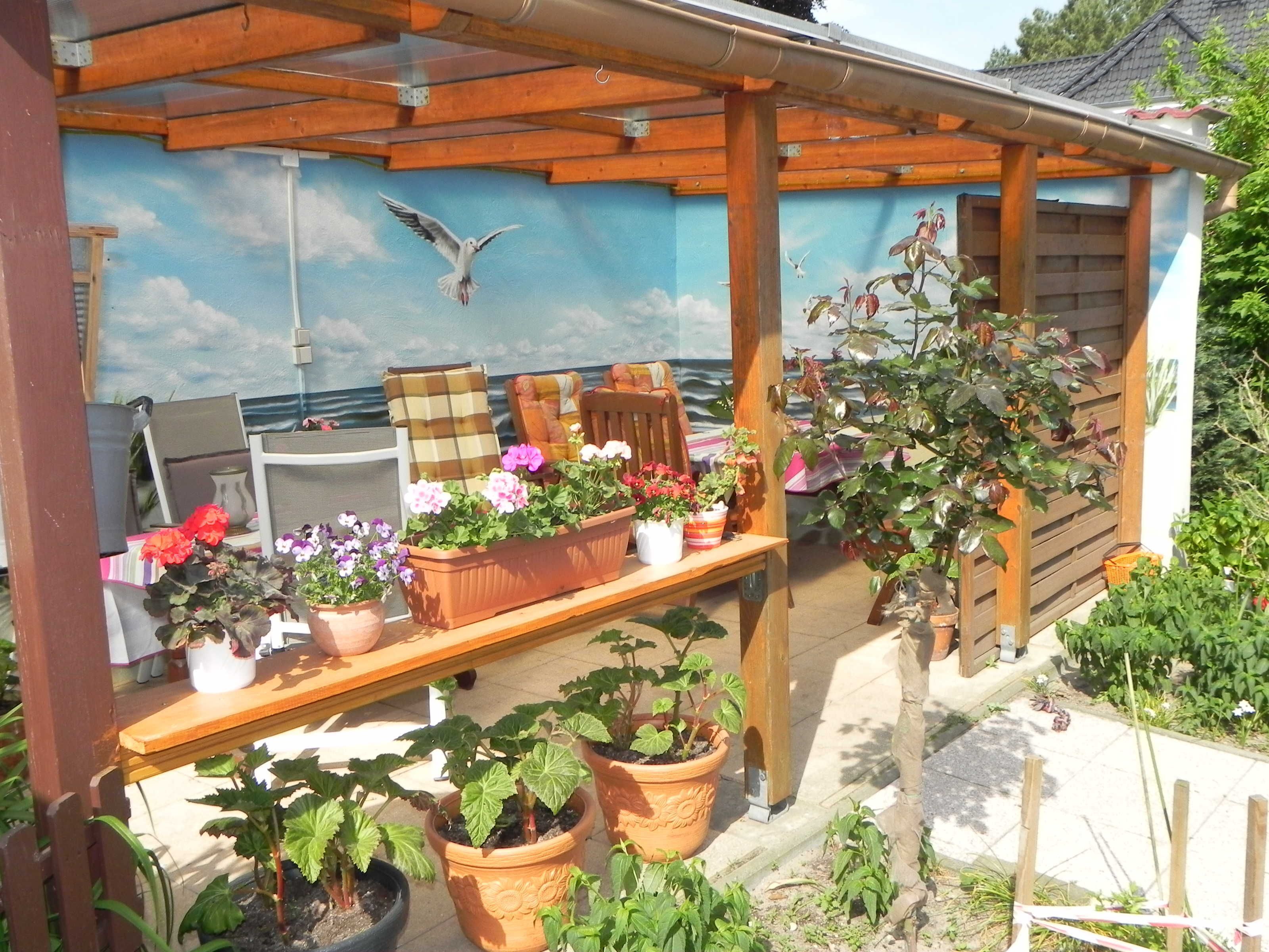 A covered outdoor seating area with a wooden bench, potted plants, and a mural of a seascape with a bird.