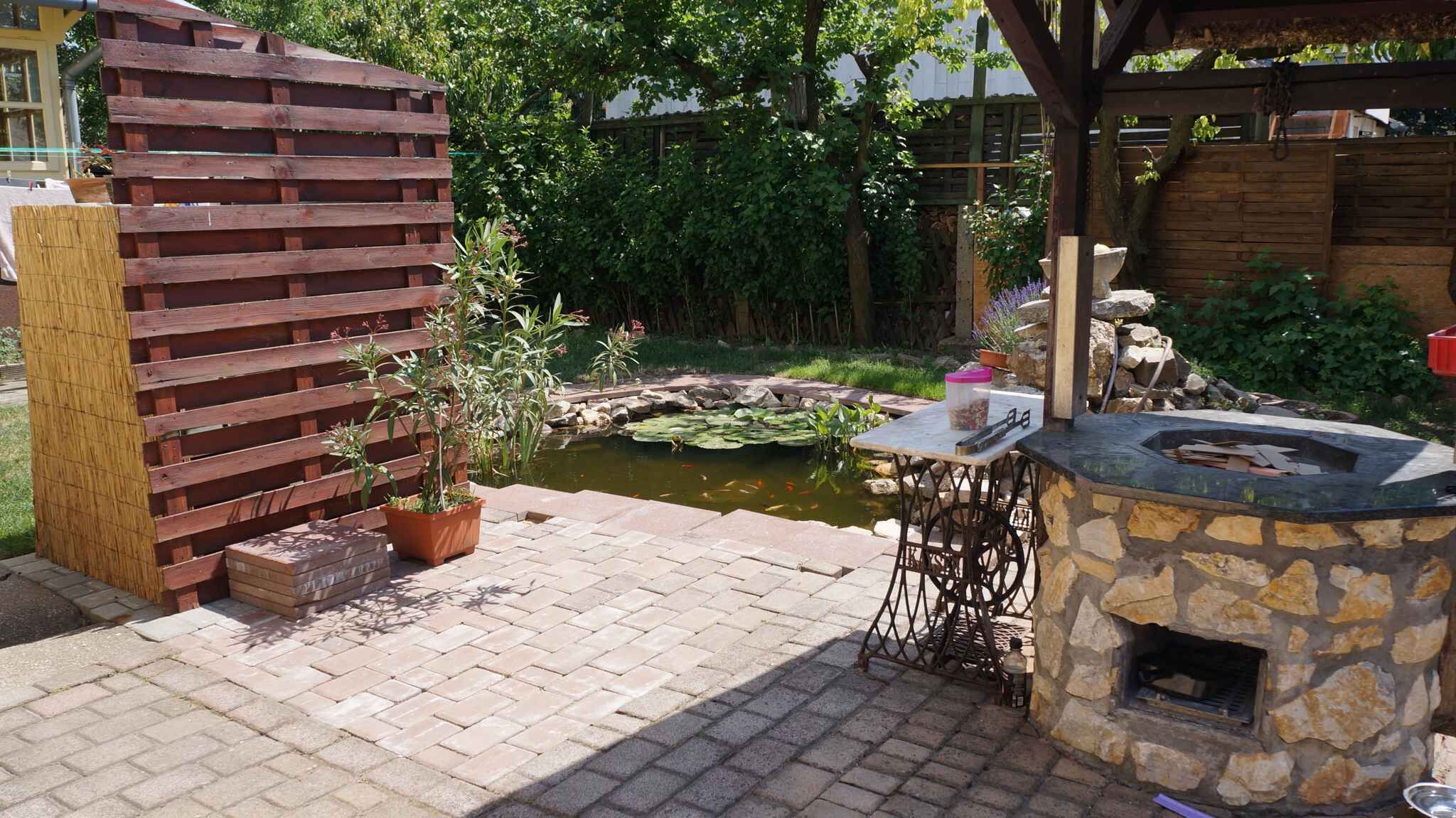 A patio area with a stone barbecue grill, a small pond, and a wooden fence.