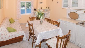 A cozy dining area with a table set for four, surrounded by wooden chairs. A floral sofa with green cushions is positioned to the left. The room features a window with a view of a body of water and trees, a small shelf with decorative items, and a kitchen area with a sink and cabinets. - Stunning home in Vela Luka with kitchen (Vela Luka)