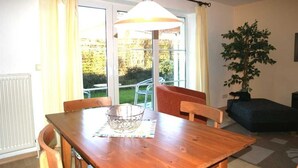 A dining area with a wooden table, chairs, a hanging lamp, and a view of the outdoors through large windows.