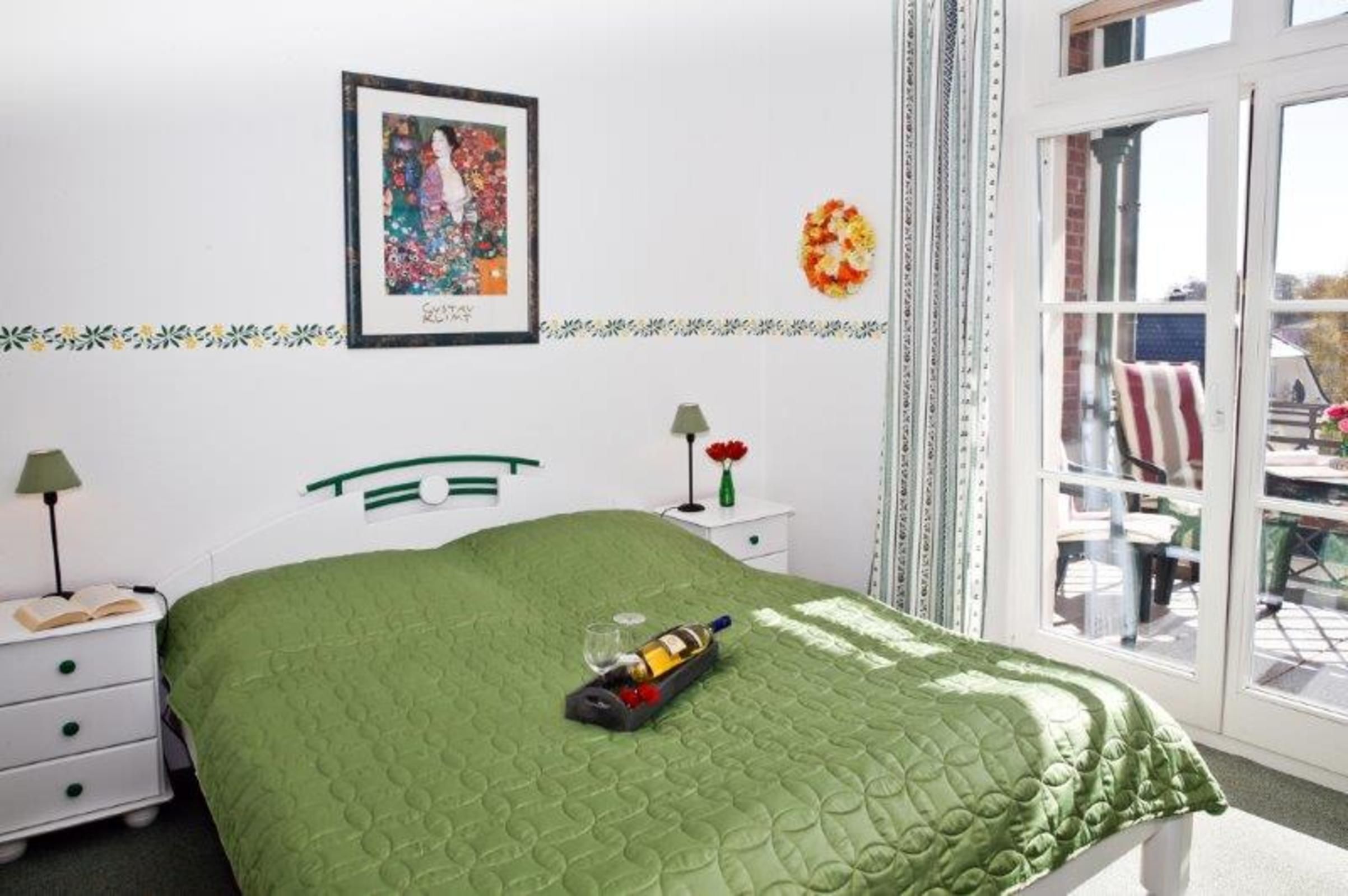 A bedroom with a green quilted bedspread, a framed picture on the wall, a small bedside table with a lamp, and a window with a view of the outside.