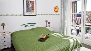 Een slaapkamer met een groen gewatteerde spreideken, een ingelijste foto aan de muur, een klein nachtkastje met een lamp en een raam met uitzicht op het buiten.