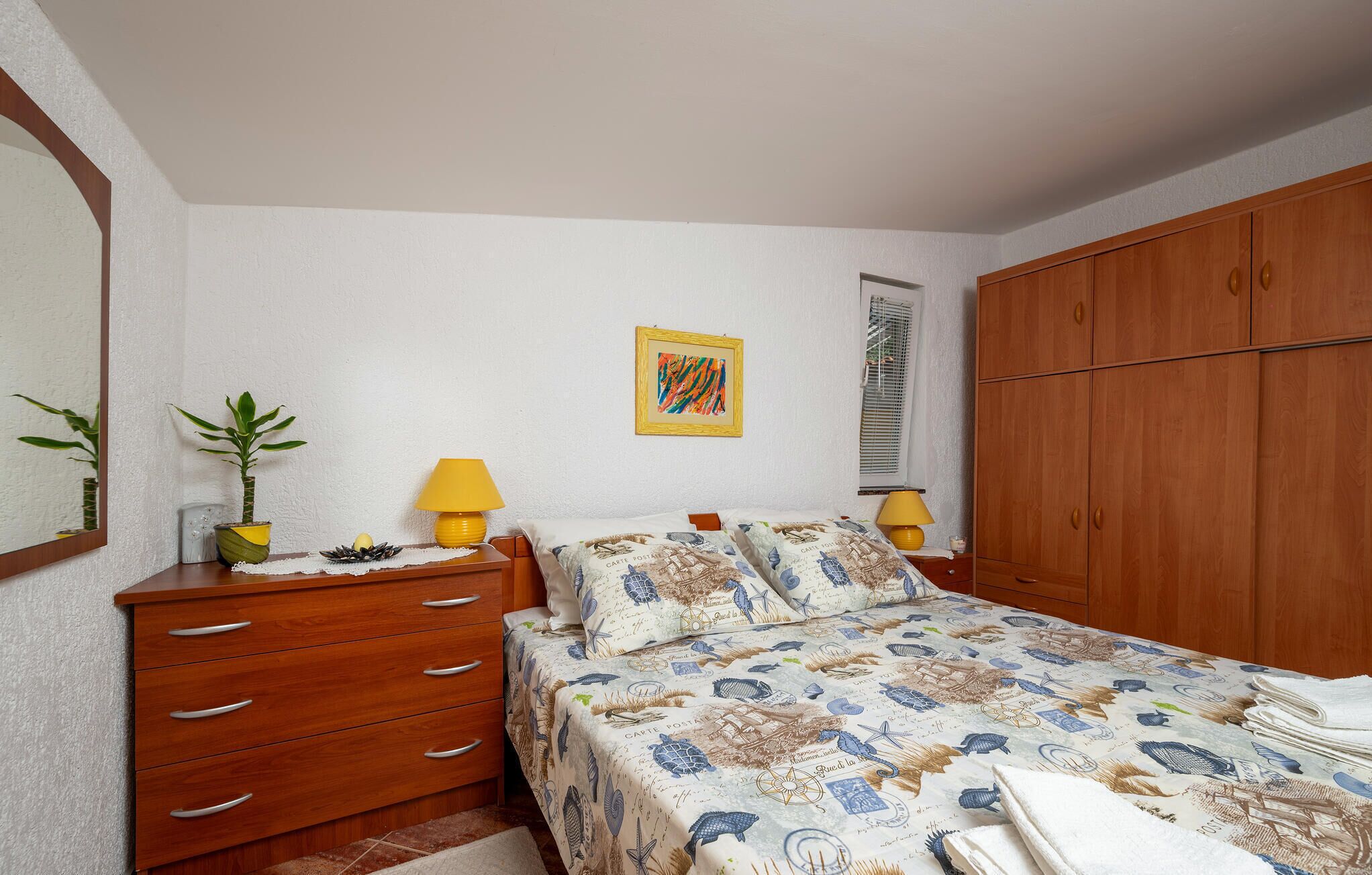 Un dormitorio con cama, armario de madera, lámparas de noche, una mesita con una planta y un cuadro en la pared.