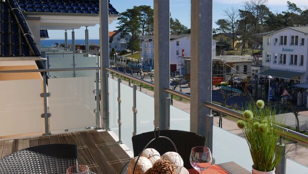 Un balcon avec une table dressée pour deux personnes, agrémentée d’une assiette de pâtisseries et de verres à vin, donnant sur une ville avec ses bâtiments et un ciel dégagé.