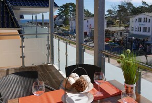 Un balcón con una mesa dispuesta para dos personas, que incluye un plato de pasteles y copas de vino, con vistas a un pueblo con edificios y un cielo despejado.