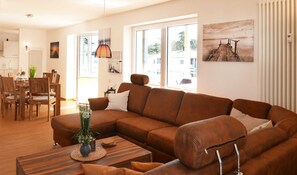 Un salon moderne avec un canapé d’angle marron, une table à manger avec des chaises et une grande fenêtre laissant entrer la lumière naturelle.