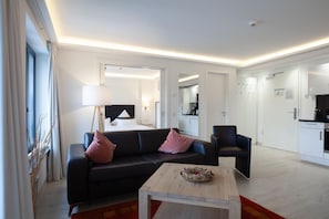 Una sala moderna con sofá, una mesa de centro y una silla. Se ve un área de cocina a través de una puerta abierta.